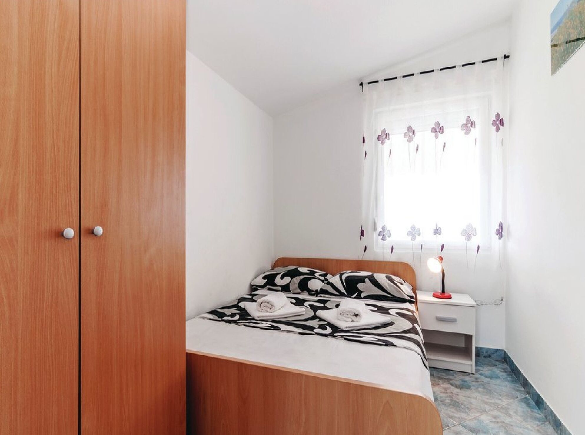 A bedroom with a wooden wardrobe, a bed with a patterned bedspread, a bedside table with a lamp, and a window with floral curtains.