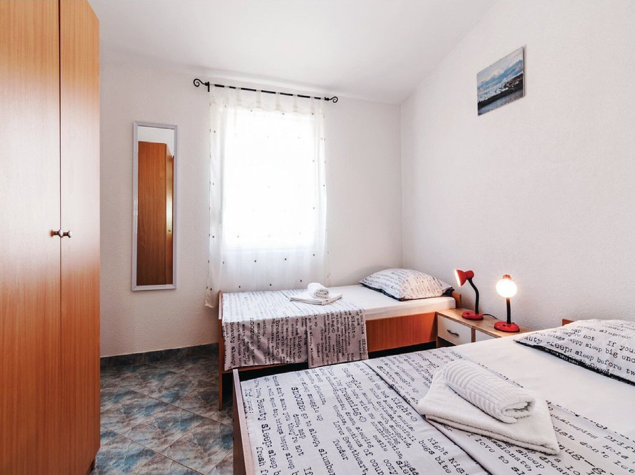 A bedroom with two beds, a window with sheer curtains, a bedside table with a lamp, and a mirror on the wall.