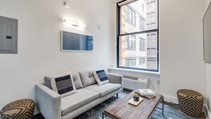 Un salon moderne avec un canapé gris, une table basse en bois et des poufs à motifs. Une grande fenêtre offre une vue sur les bâtiments à l’extérieur.