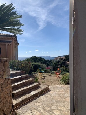 Una escalera de piedra conduce a una terraza con vistas a un paisaje costero, que incluye un edificio con tejado de baldosas y una palmera.
