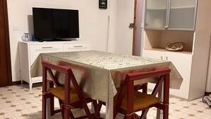 Zona pranzo con tavolo ricoperto da tovaglia, circondato da quattro sedie con cuscini. Un televisore è montato su un mobile bianco, e un cesto è posizionato su uno scaffale del mobile.
