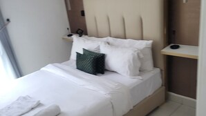 Ein ordentlich bezogenes Bett mit weißen Leinen und einem grünen Kopfkissen, ein kleiner Nachttisch mit einer Lampe und ein an der Wand befestigtes Regal.