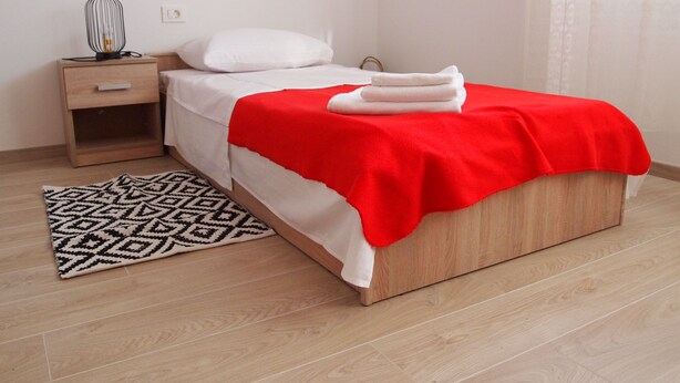 Ein ordentlich bezogenes Bett mit roter Decke, ein Nachttisch mit Lampe und ein gemustertes Teppich auf Holzboden.