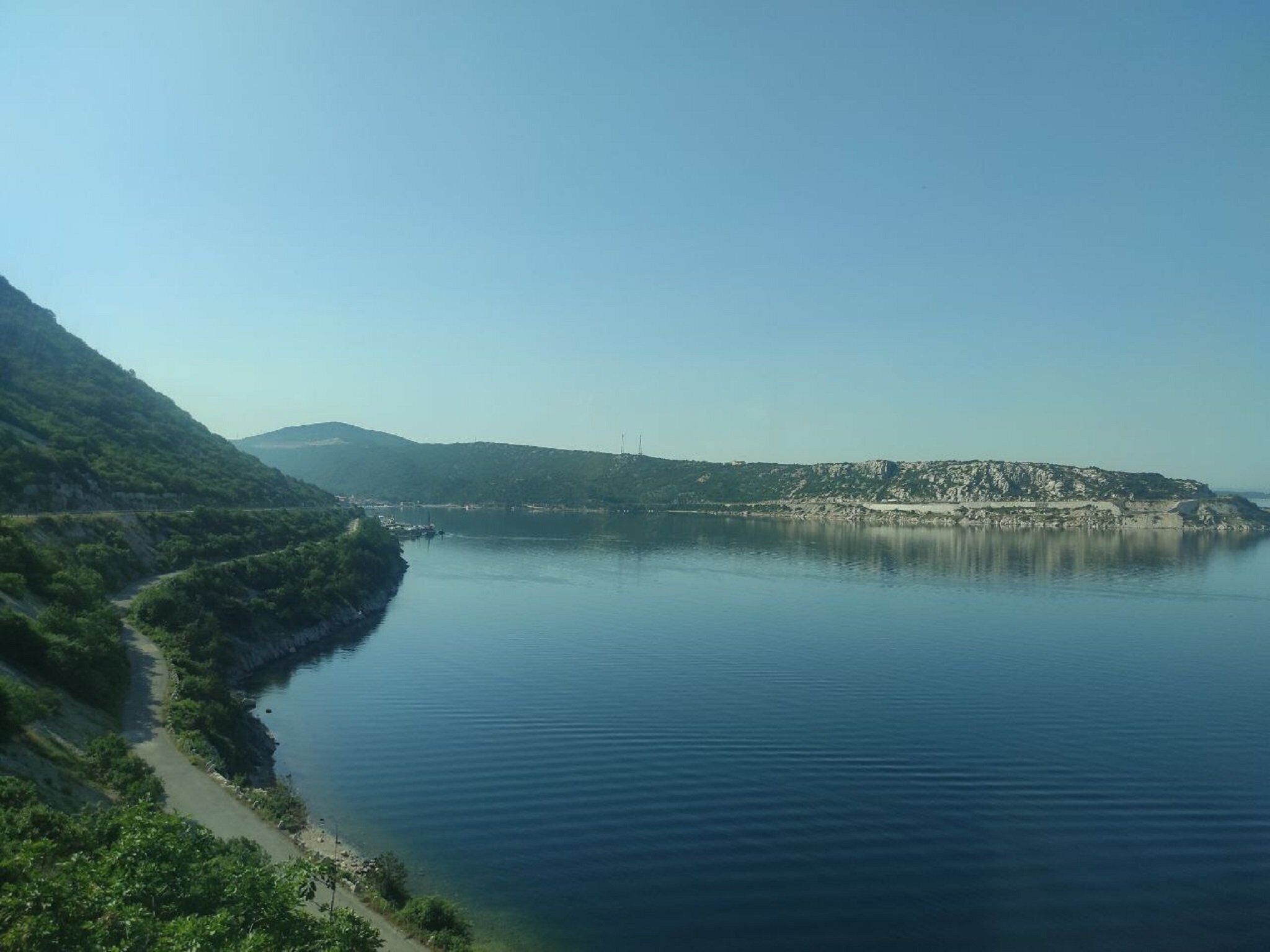 A calm lake with a narrow path along the shore, surrounded by hills.