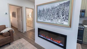 Uma sala de estar moderna com lareira, uma fotografia emoldurada de uma floresta no inverno, e uma área de cozinha ao fundo.