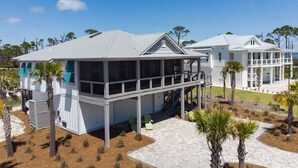 Een huis aan de kust met een overdekte veranda, een balkon op de tweede verdieping, en een aangelegde tuin met palmboompjes.