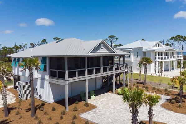 A coastal house with a covered porch, a second-story balcony, and a landscaped yard with palm trees.