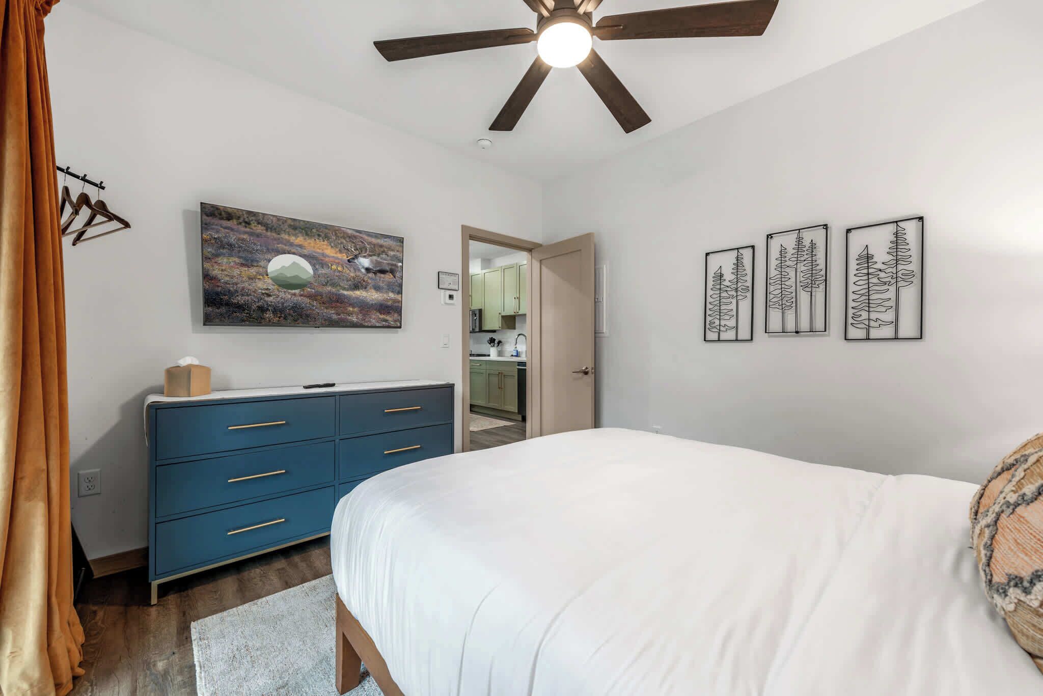 A bedroom with a bed, a dresser, a ceiling fan, and artwork on the walls.