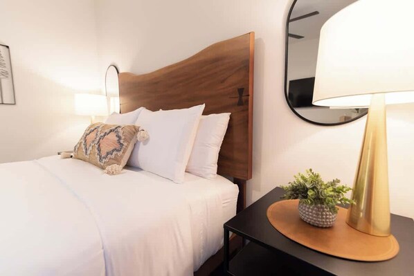 Una cama bien tendida con sábanas blancas y un cojín decorativo estampado, una mesita de noche con una lámpara y una planta pequeña, y un espejo instalado en la pared.