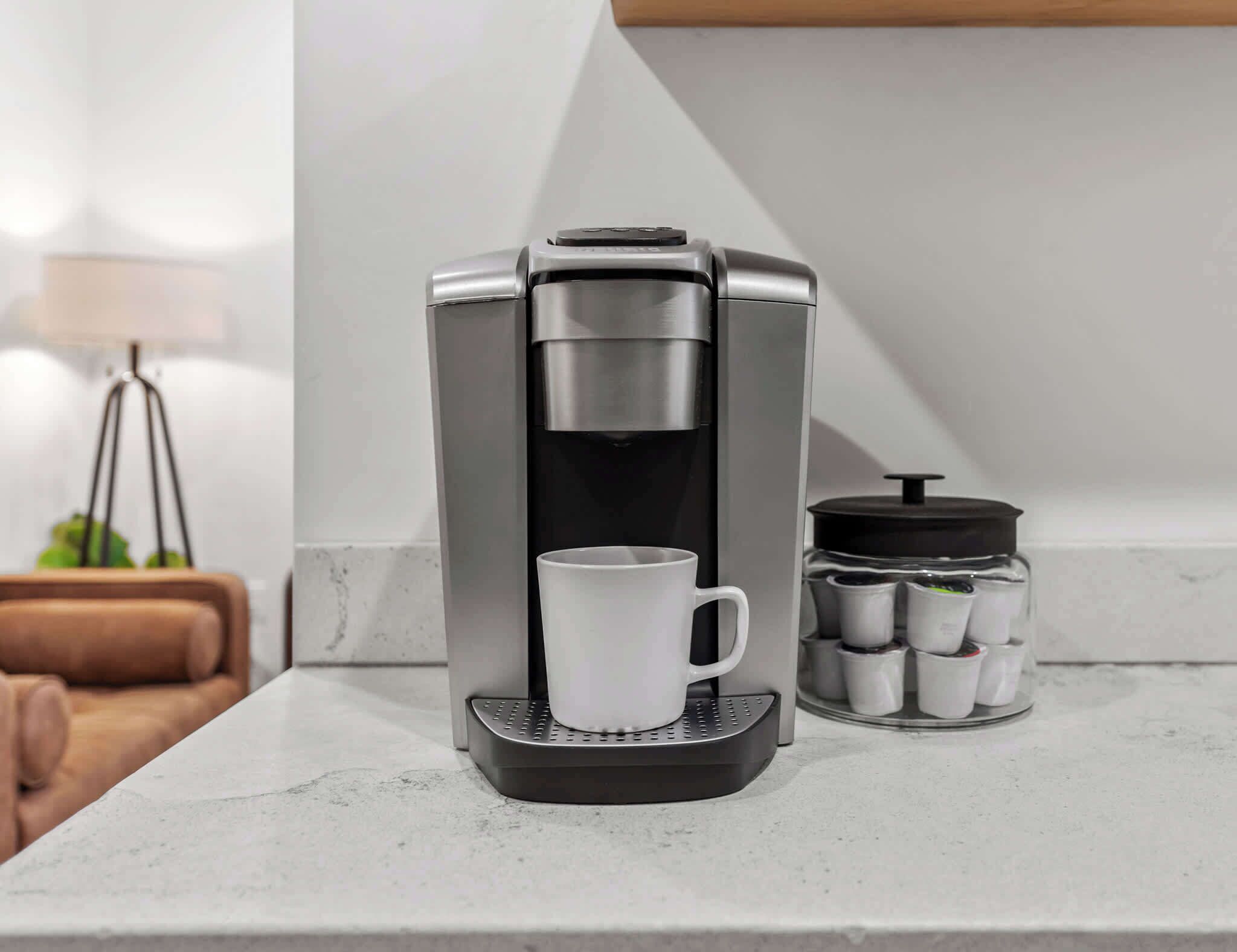 A coffee maker with a white mug on a countertop, a small container with tea bags, and a lamp in the background.