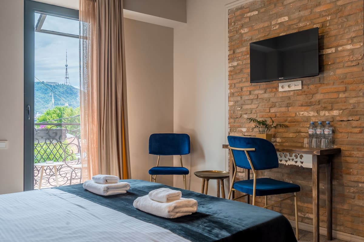 Hotelkamer met een bed, twee blauwe stoelen, een klein tafeltje, een aan een bakstenen muur bevestigde televisie, en uitzicht op groen buiten.