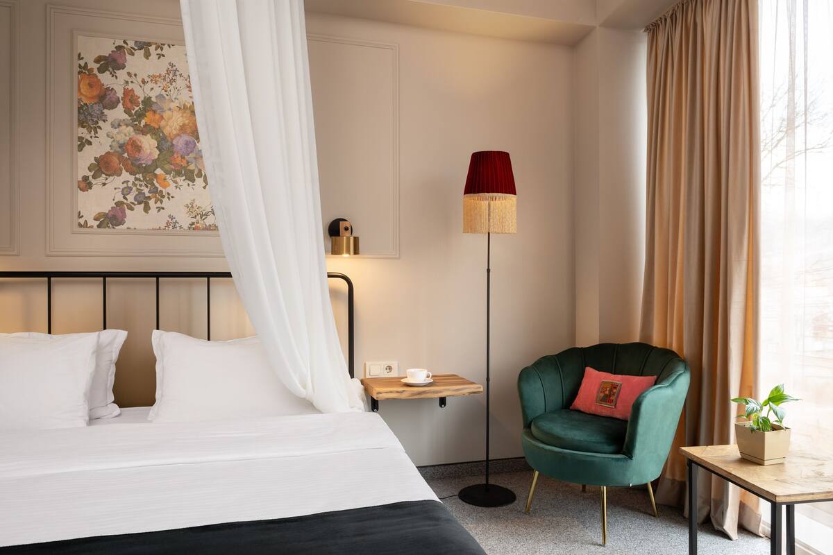 Een netjes ingerichte hotelkamer met een bed, een nachtkastje met een kop, een groene fauteuil, een vloerlamp en een aan de muur hangend schilderij.