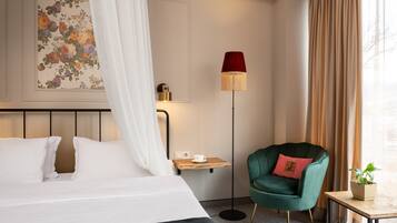 A neatly arranged hotel room with a bed, a bedside table with a cup, a green armchair, a floor lamp, and a wall-mounted painting.