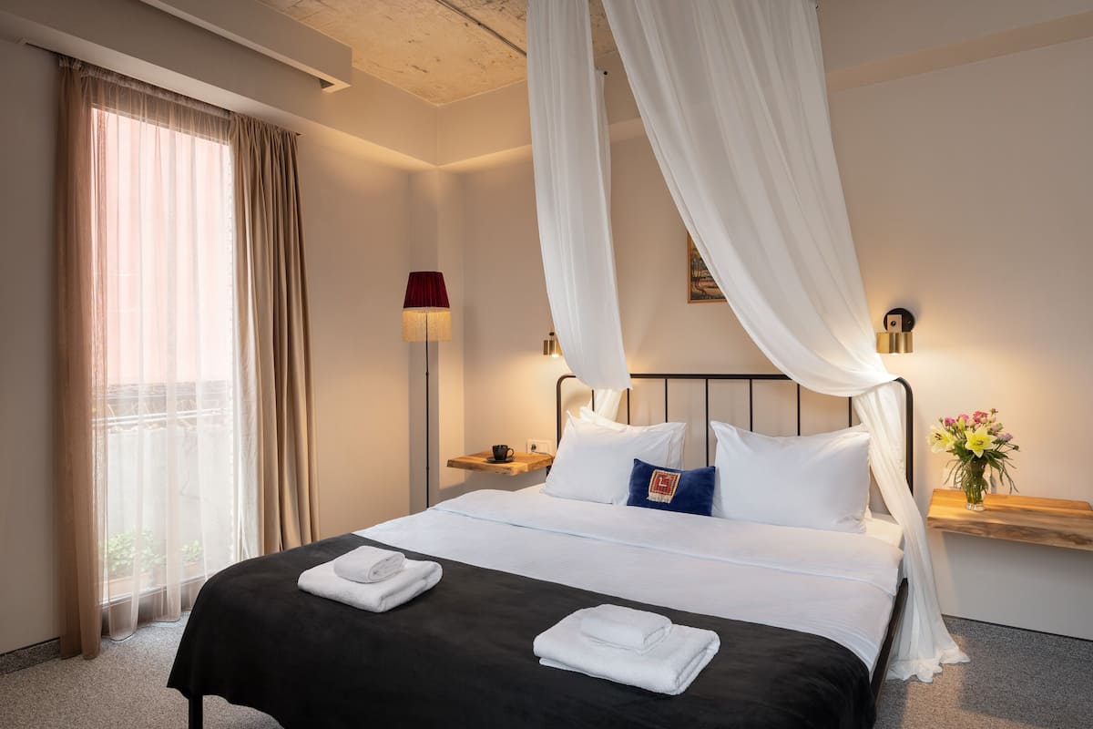 Een net opgemaakt bed met witte lakens, een nachtkastje met een lamp, en een raam met dunne gordijnen.