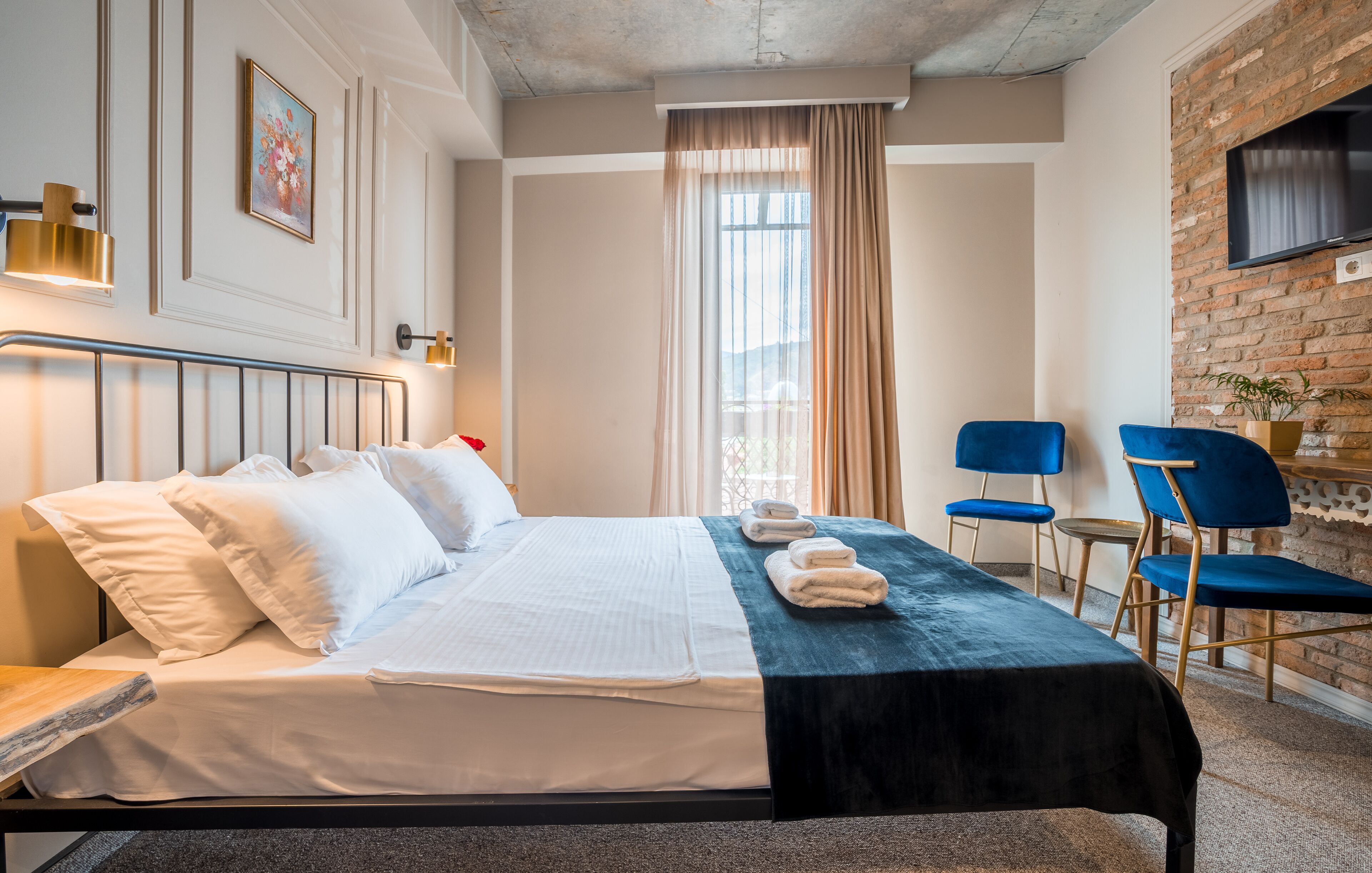 A hotel room with a bed, a desk, two chairs, a TV, and a window with curtains.
