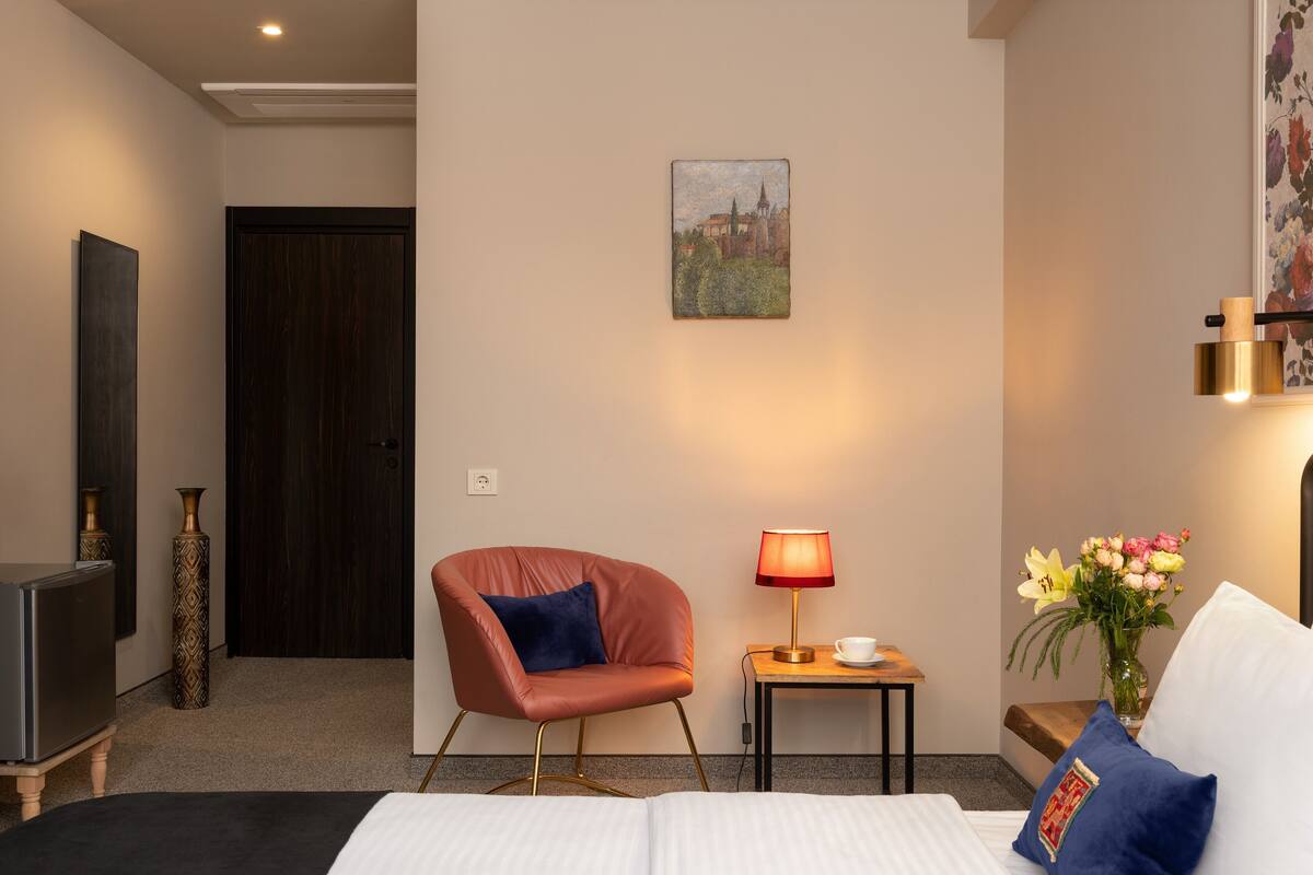 Een hotelkamer met een bed, een stoel, een televisie, een lamp, een bijzettafeltje en een vaas met bloemen.