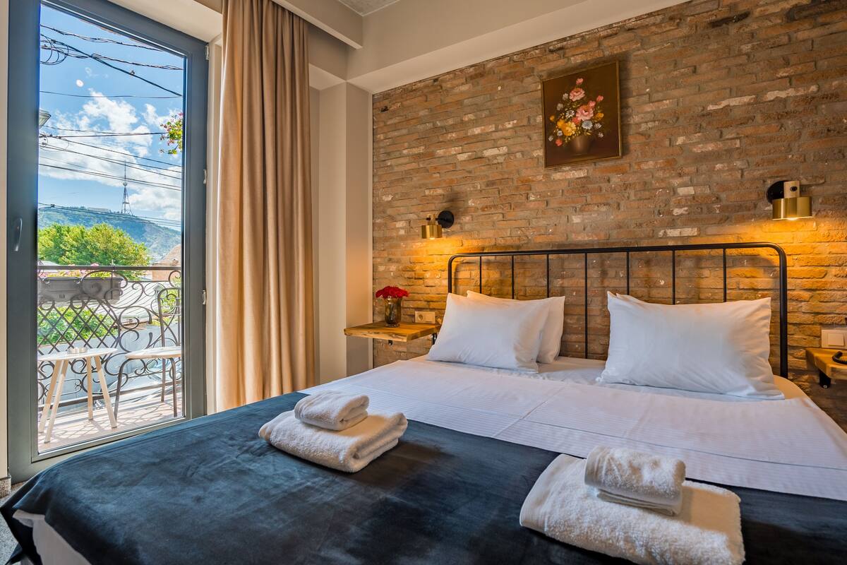 Hotelkamer met een bakstenen muur, een groot bed met witte lakens, een balkon met uitzicht en een schilderij boven het bed.