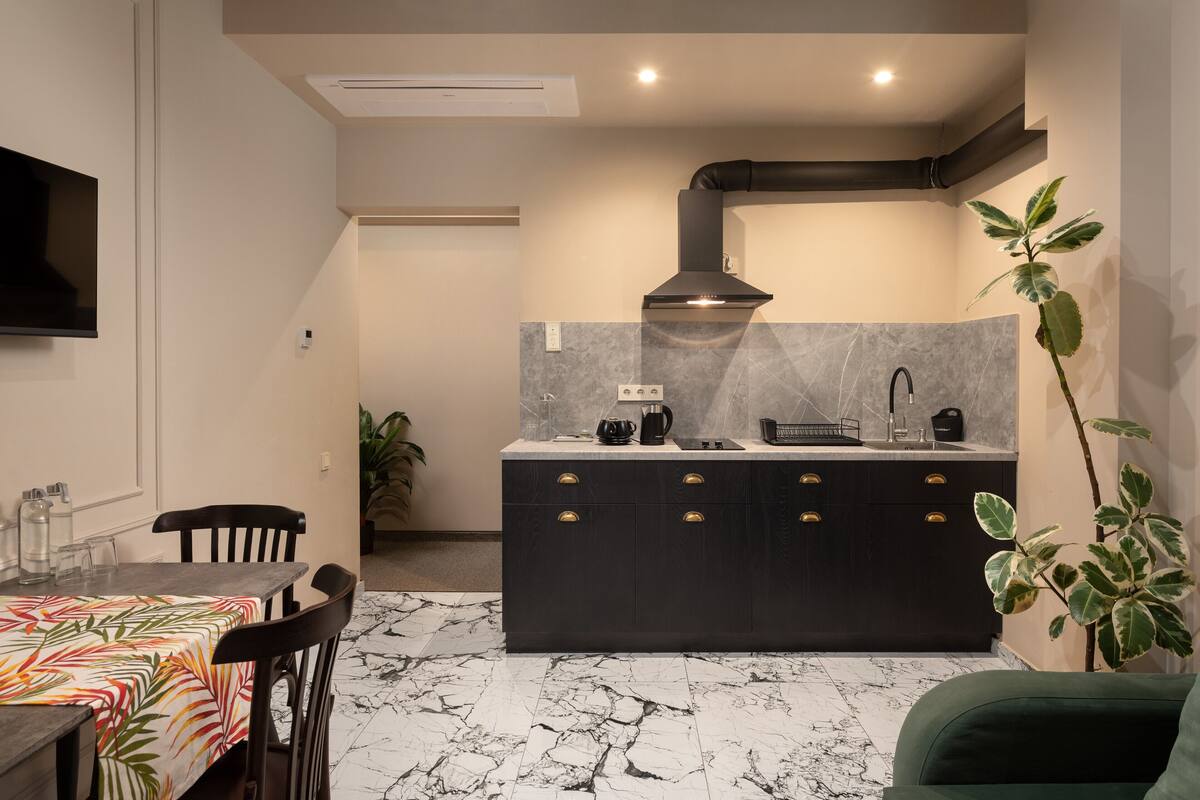 Een moderne keuken met een zwart aanrecht, een marmeren spatwand en een zwarte afzuigkap. Er is een eettafel met stoelen en een groene bank in de woonkamer.