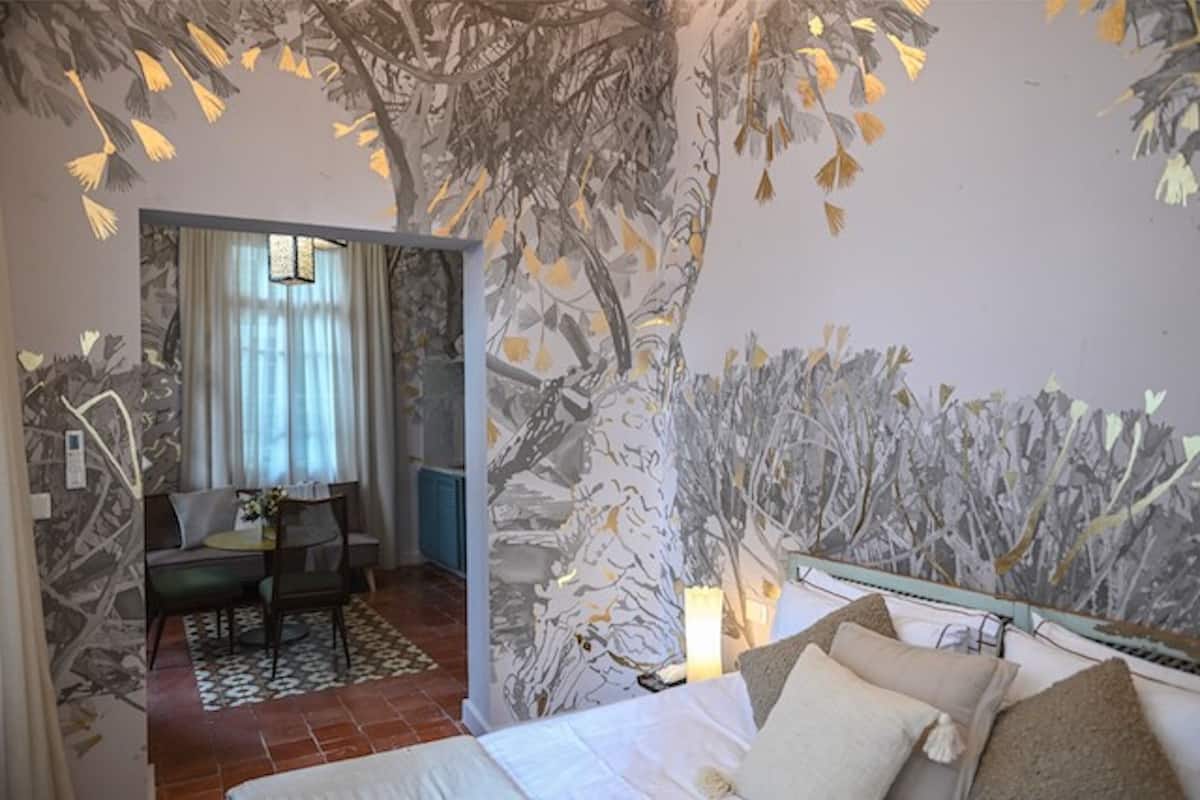 A bedroom with a bed, a desk, and a chair. There is a window with curtains and a lamp on the desk.