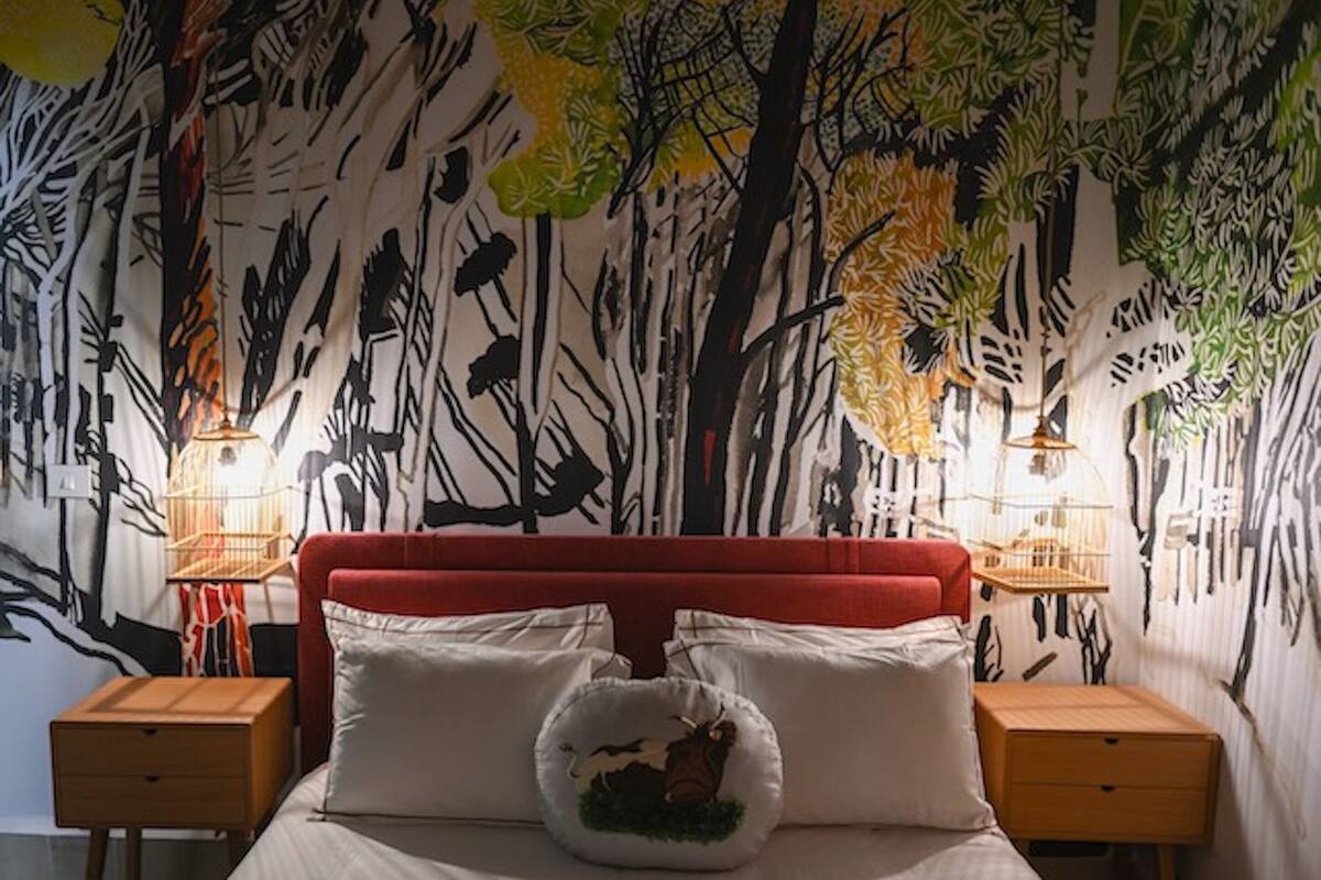 A bedroom with a red upholstered headboard, two bedside tables with lamps, and a wall mural of trees and leaves.