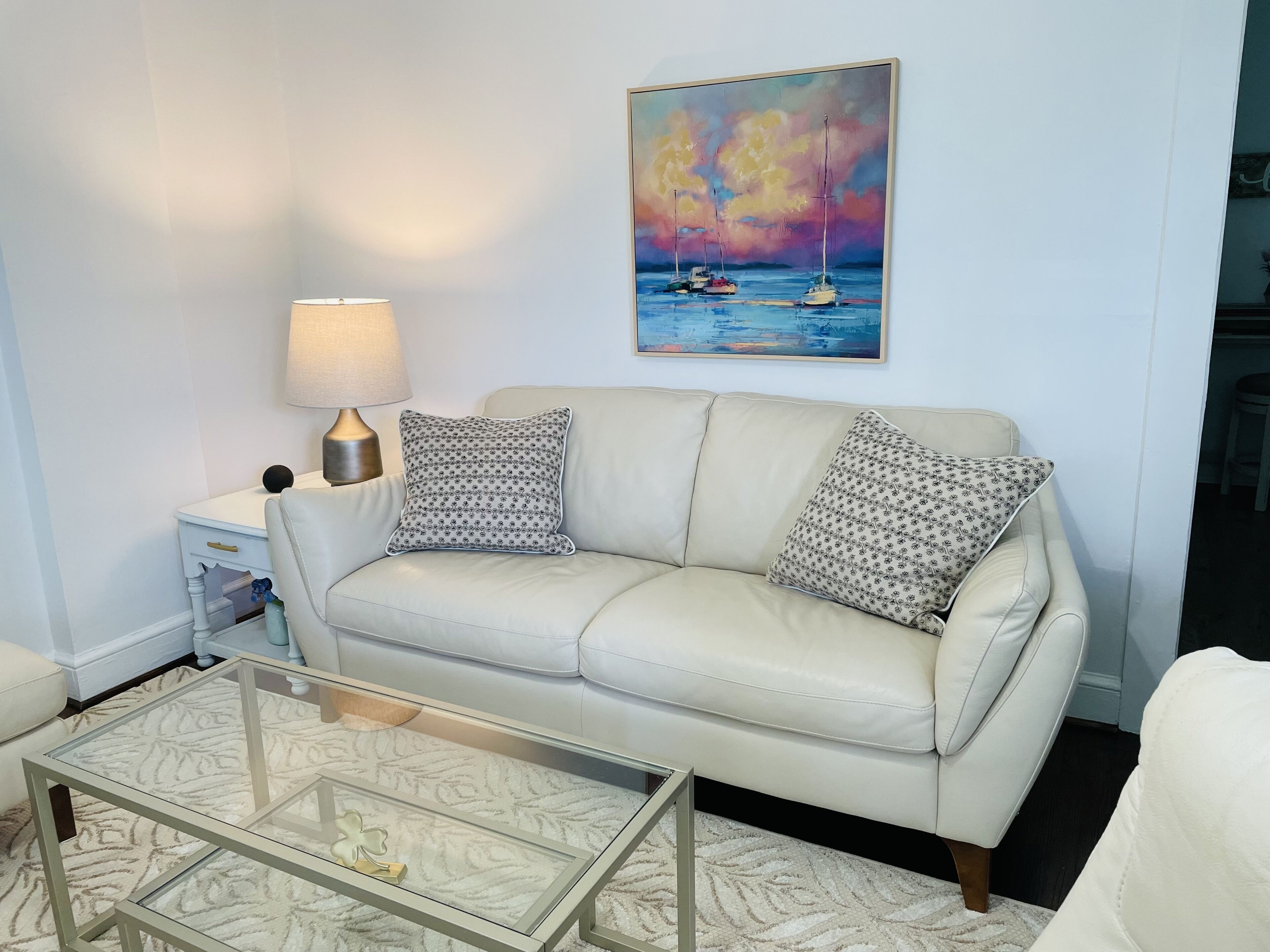 A living room with a white leather sofa, two patterned pillows, a glass coffee table, a lamp, and a painting of a seascape.