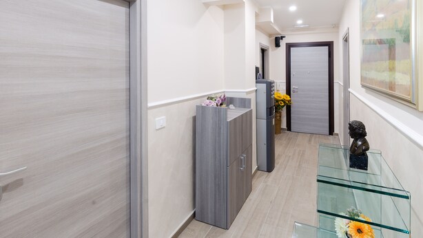A hallway with a wooden door, a grey cabinet, and a glass table with a sculpture and flowers. - AFFITTACAMERE ALESSIA (Sanremo)