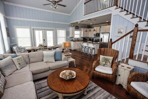 A spacious living room with a large sofa, a round coffee table, and a wicker armchair. A kitchen is visible in the background with white cabinets and stainless steel appliances.