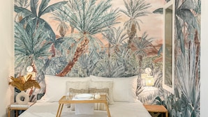 A bedroom with a large mural of tropical foliage, a bed with a white textured bedspread, and a bedside table with a vase and books. - Americana beach house (Rimini)