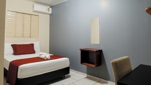 A hotel room with a bed, a small table, a chair, a wall-mounted shelf, and an air conditioning unit.