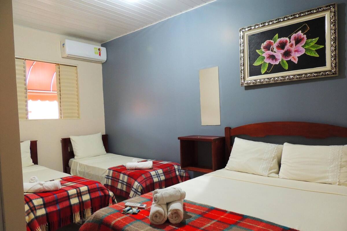 A hotel room with two beds, a red and blue checkered blanket on one bed, a painting of flowers on the wall, and an air conditioning unit.