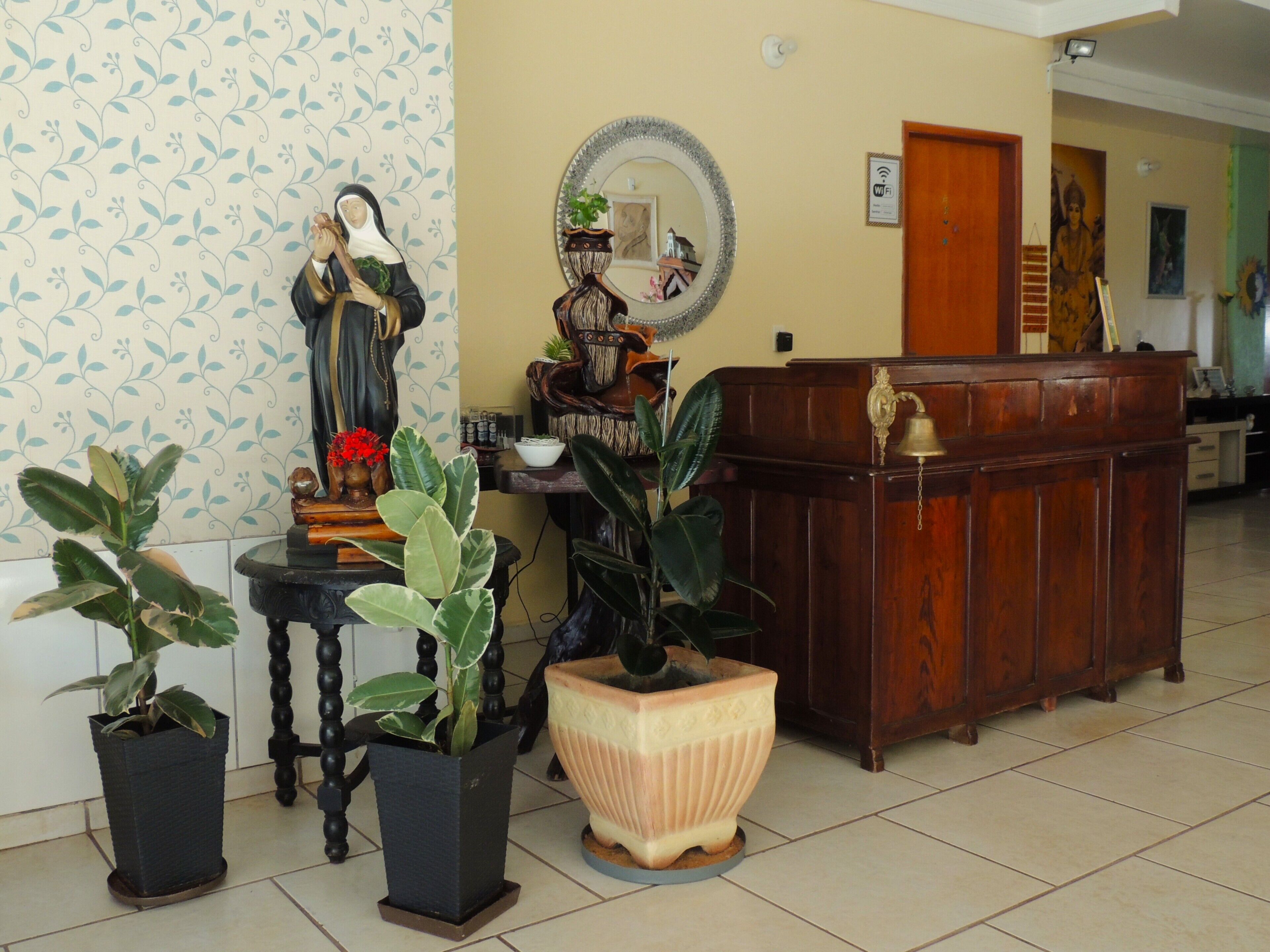 Um quarto com uma mesa de recepção de madeira, uma estátua, plantas em vasos e um espelho na parede.