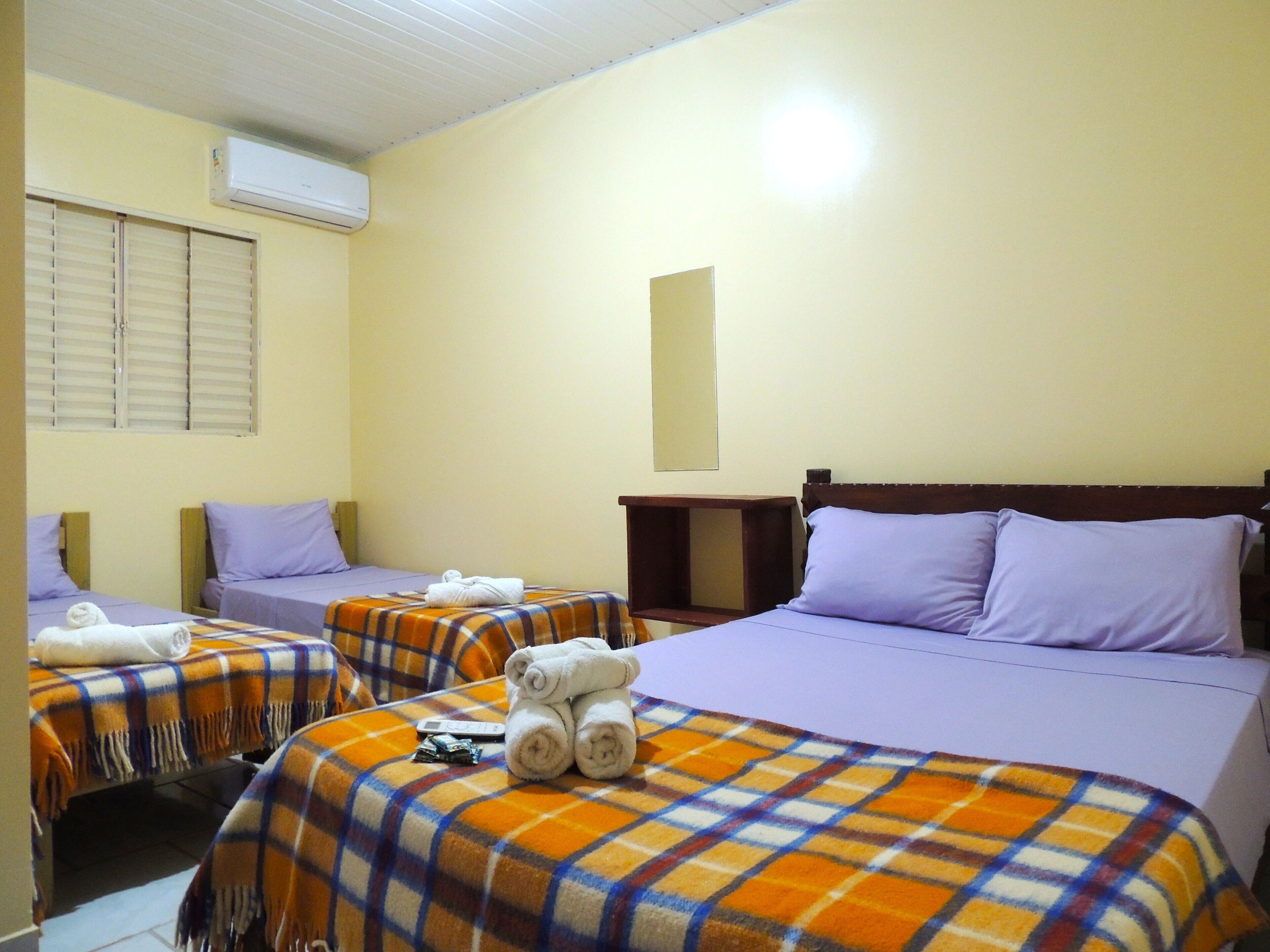 Una habitación de hotel con dos camas, cada una con un almohadón morado y una manta escocesa. Hay una mesita de noche de madera con una lámpara y un equipo de aire acondicionado en la pared.
