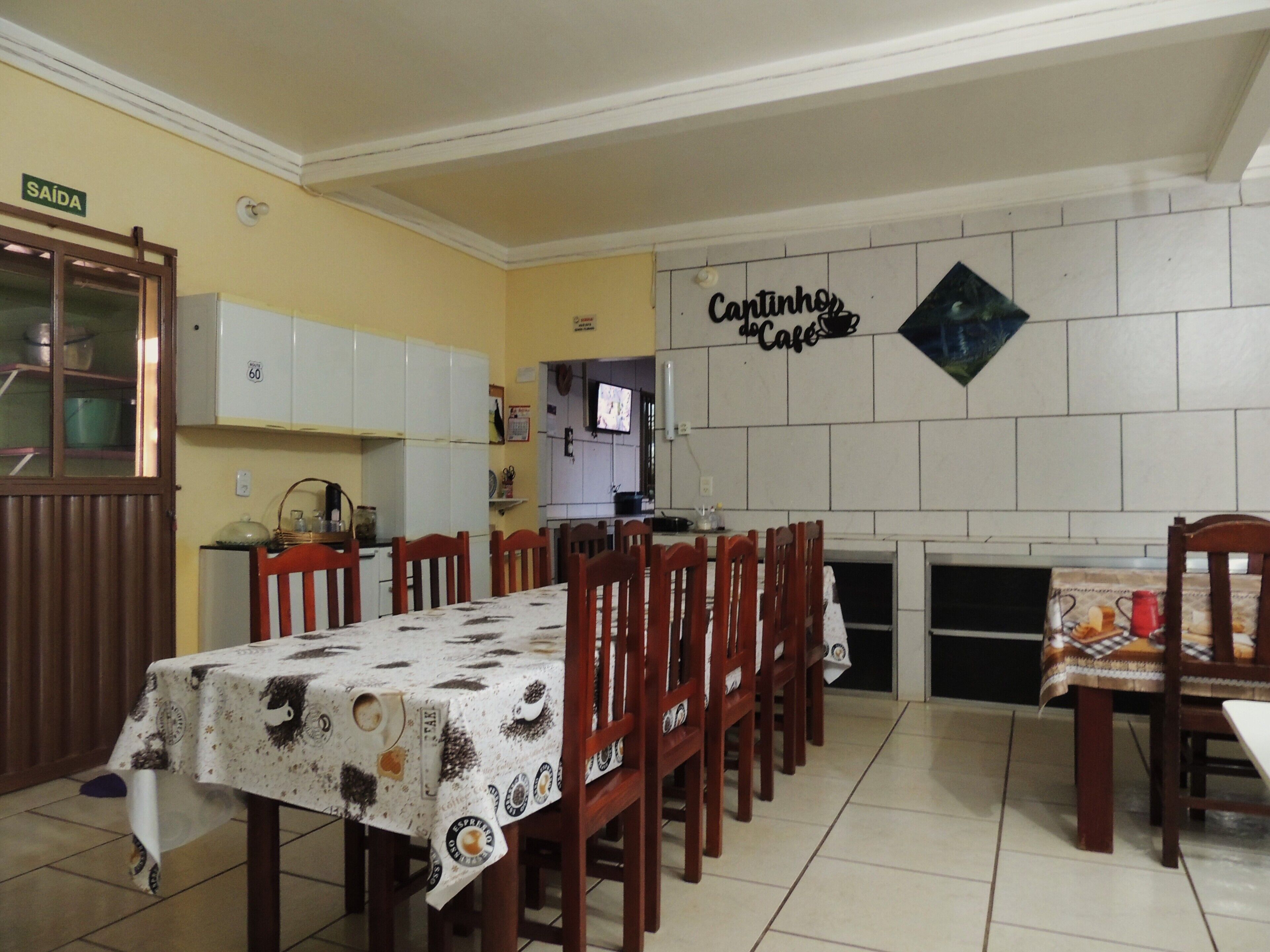 Comedor con una mesa dispuesta para seis comensales, sillas de madera, zona de cocina con armarios y un letrero en la pared que dice 'Cantinho do Cafe'.