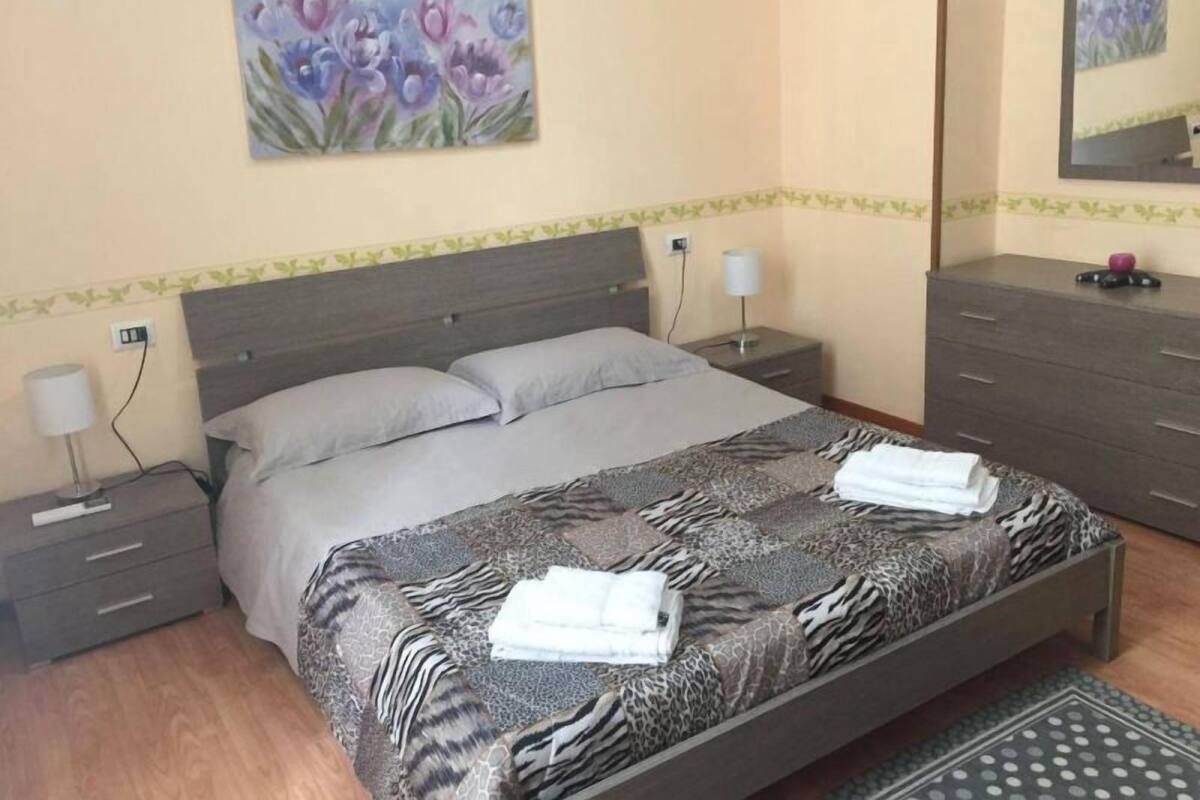 A bedroom with a zebra-patterned bedspread, a nightstand with a lamp, a dresser, and a painting of flowers on the wall.