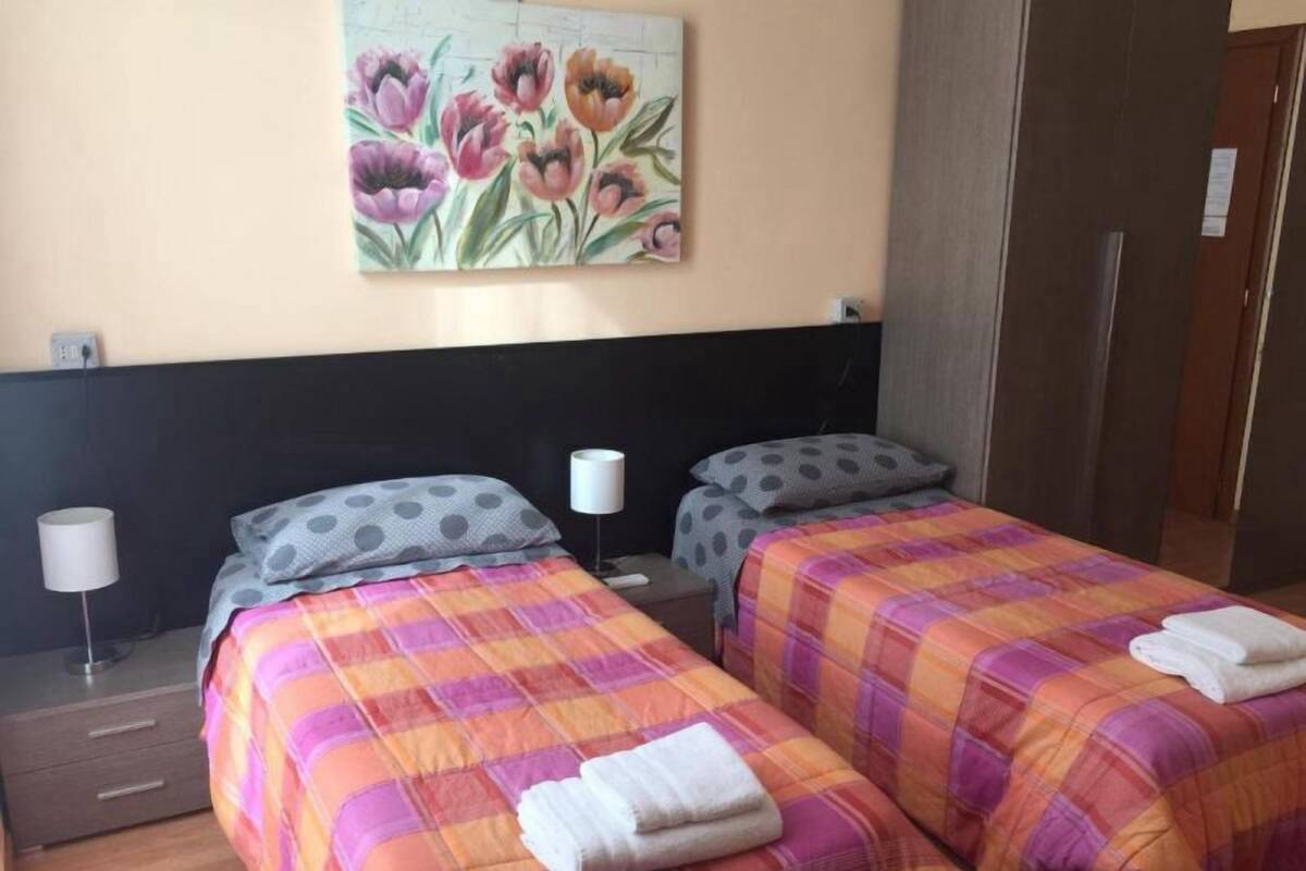 A hotel room with two beds, each with a plaid bedspread and pillows, a nightstand with a lamp, and a painting of flowers on the wall.