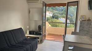 Ein Balkon mit Blick auf einen Hügel und ein Gebäude, eine schwarze Couch, ein kleiner Tisch und ein an der Wand montierter Fernseher.