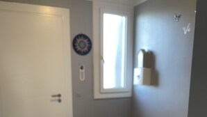 Ein kleines Zimmer mit einem Einzelbett, Holzboden, einer weißen Tür, einem Fenster mit Jalousien und einer Wandleuchte.
