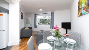 Un salon moderne comprenant un coin repas, des canapés gris, une table en verre pour quatre personnes et une grande fenêtre avec des rideaux.