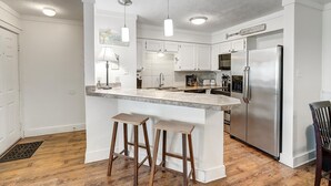 Una cucina moderna con mobili bianchi, un frigorifero in acciaio inossidabile, un piano di lavoro in marmo e due sgabelli in legno.