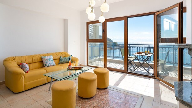 Un salón luminoso con un sofá amarillo, una mesa de centro de cristal y otomanas. Cuenta con un amplio ventanal corredera que da a un balcón con una mesa y sillas.