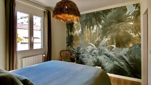 A bedroom with a bed, a window with curtains, a tropical mural, and a hanging light fixture.