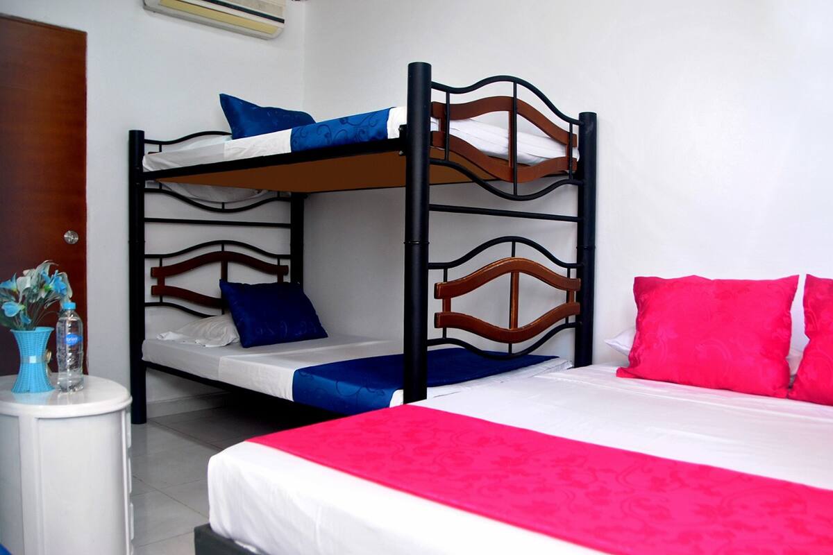 A bunk bed with two levels, a bedside table with a vase and water bottle, and a bed with a red and white bedspread.
