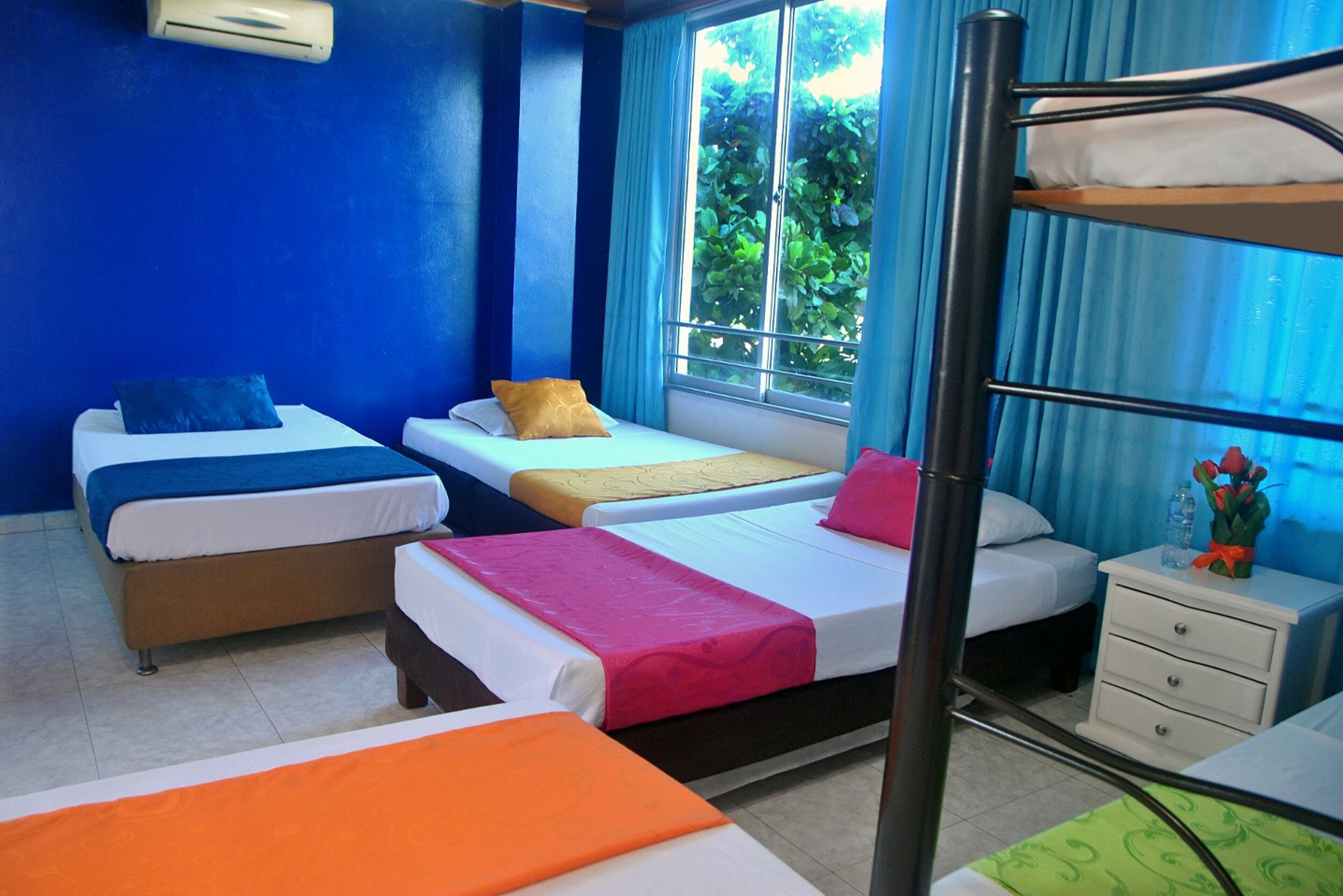 Habitación de hotel con cuatro camas, una litera, una ventana con vistas a vegetación y una mesita de noche con una botella de agua y flores.