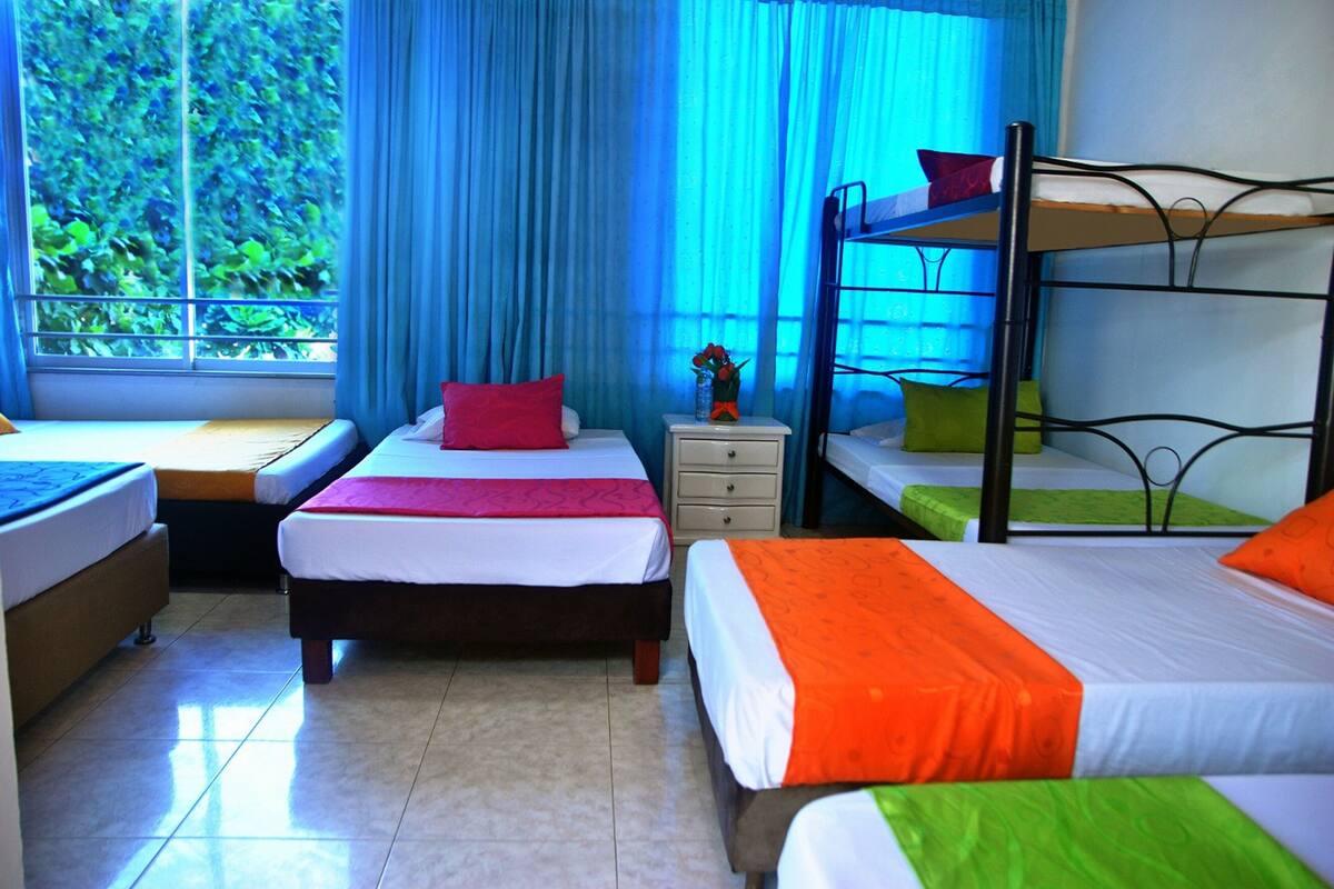 A hotel room with two single beds, a bunk bed, a nightstand with a vase of flowers, and a window with blue curtains.