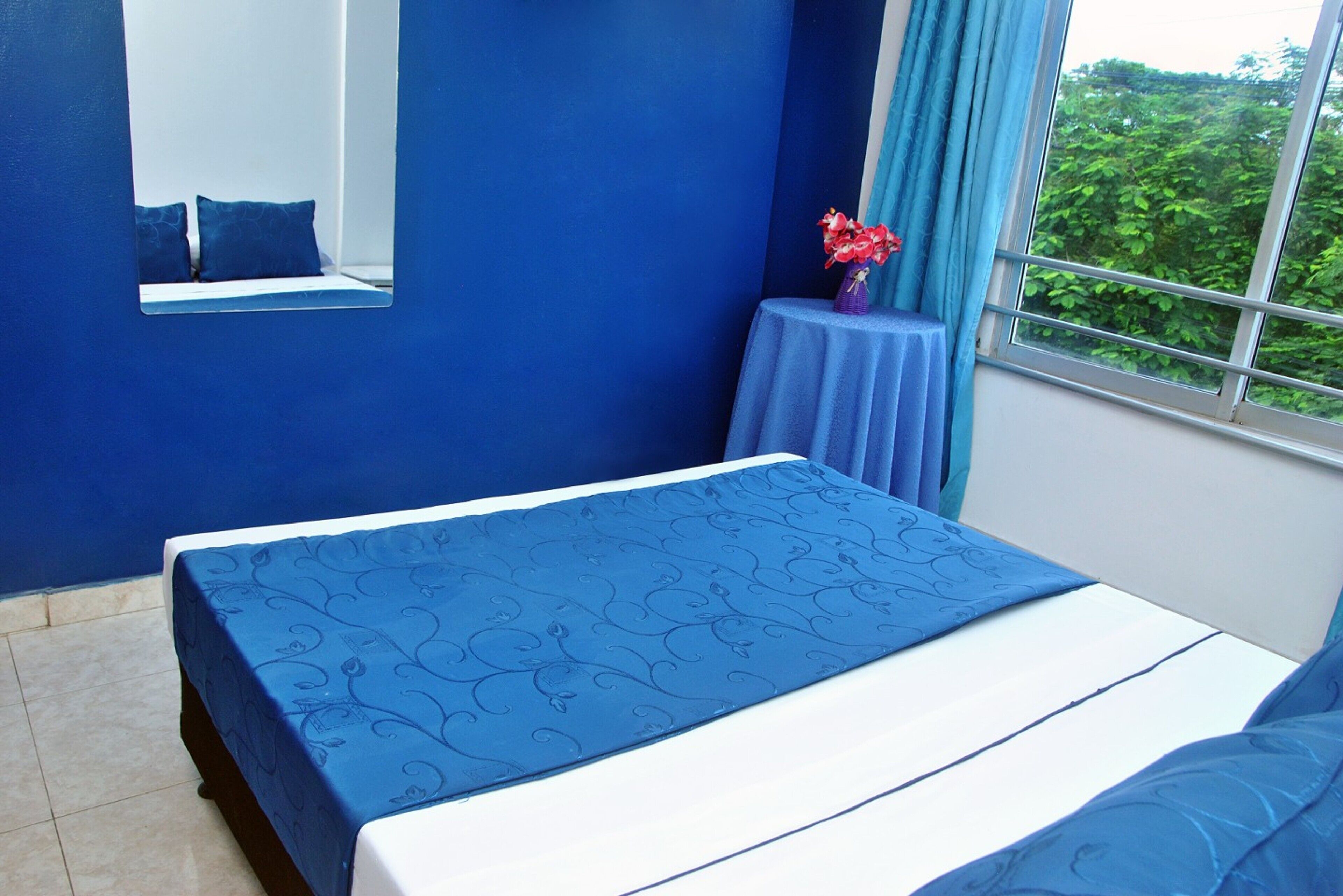 Una habitación con una cama, una mesita con un florero, un espejo y una ventana con vistas a follaje.