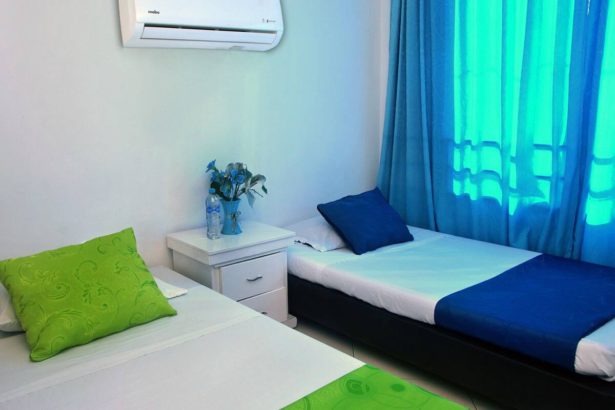 A hotel room with two beds, a nightstand, a vase with flowers, and a window with blue curtains.