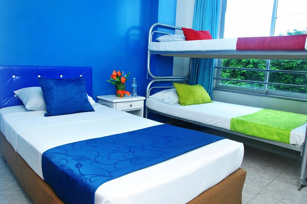 A hotel room with two beds, one with a blue headboard and the other with a metal frame. There is a window with curtains, a nightstand with a vase of flowers, and a small table.