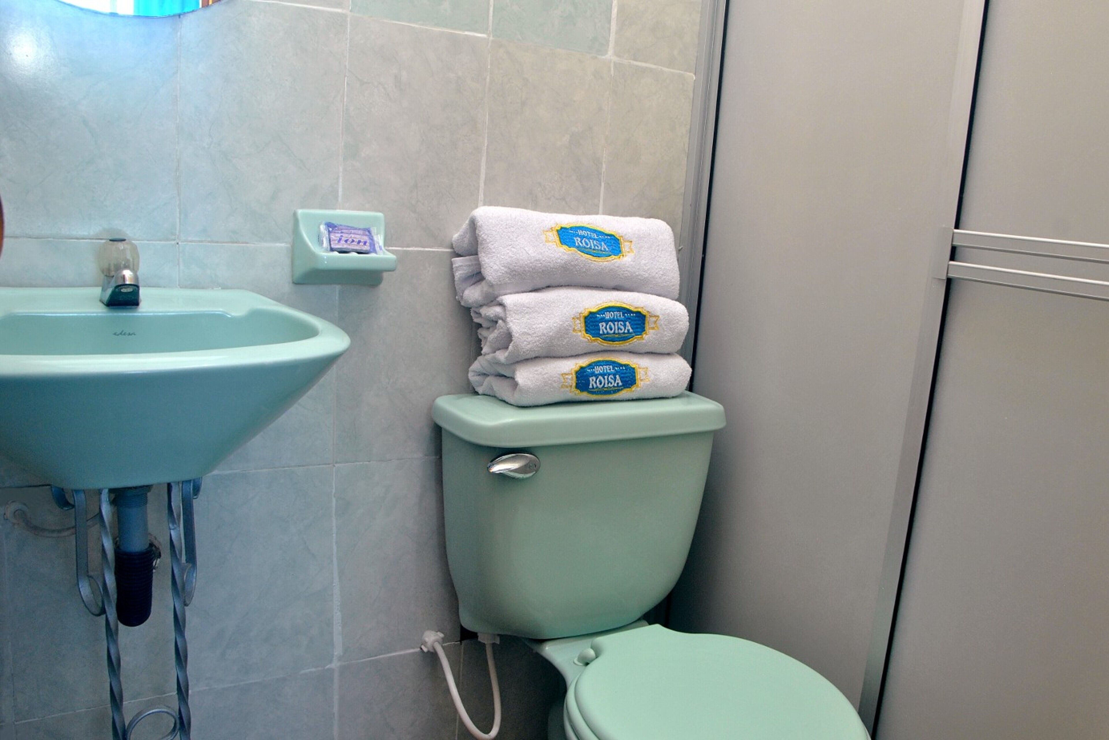 Casa de banho com lavatório verde claro, sanita e pilha de toalhas brancas com etiqueta azul.