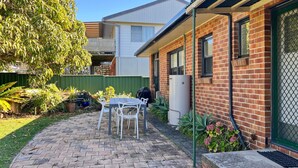 A residential backyard with a brick house, a covered patio with a table and chairs, and a variety of potted plants.