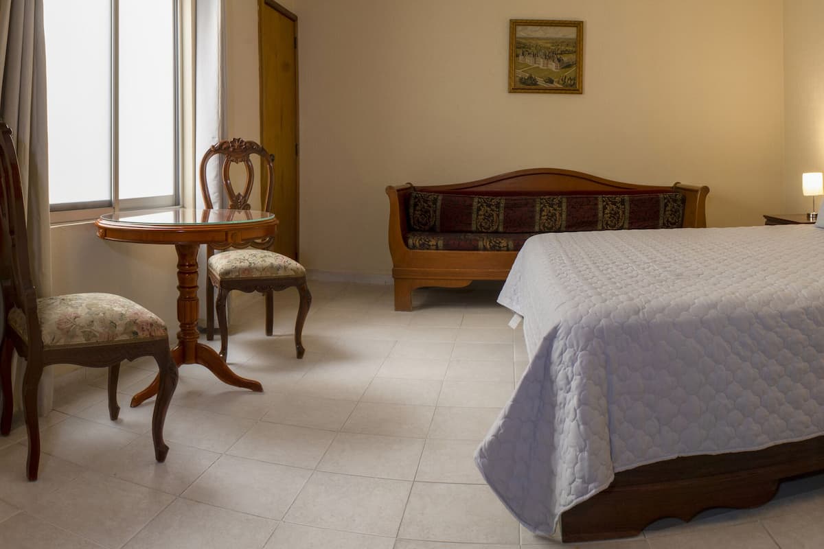 A hotel room with a large bed, a wooden nightstand, a small table with two chairs, and a painting on the wall.